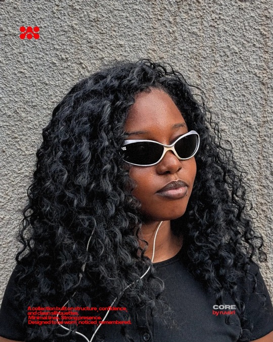 Woman with long dark curly hair and silver Ruvori sunglasses, earphones around her neck, against a textured gray wall.