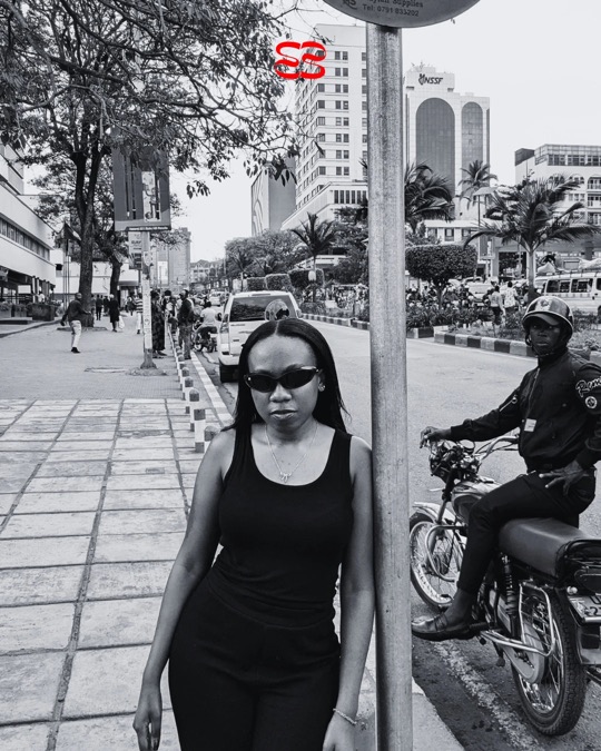A woman in black ruvori sunglasses leaning on a pole beside a busy road, in black and white.