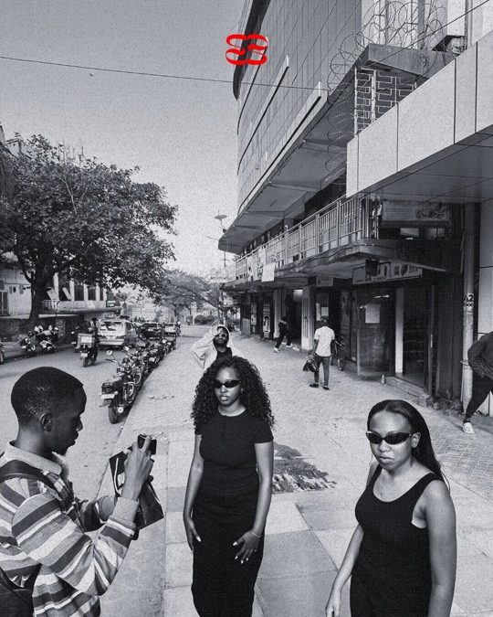 On a shoot in the street: a photographer lines up a shot of two women in black wearing ruvori sunglasses, in black and white.
